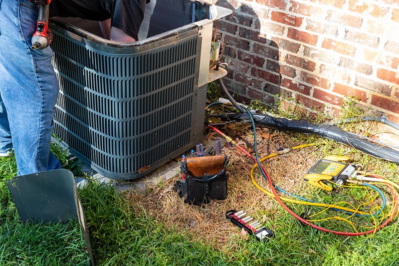 3 Tips to Prevent AC Breakdown During a Heat Wave. Photo of an air conditioning unit installed outside of a brick home being worked on by a technician.
