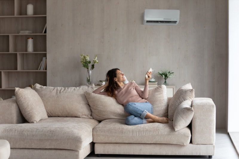 Planning to Remodel? Go Ductless. Relaxed young hispanic female homeowner sitting on huge comfortable couch, turning on air conditioner with remote controller, switching on cooler system, setting comfortable temperature in living room.